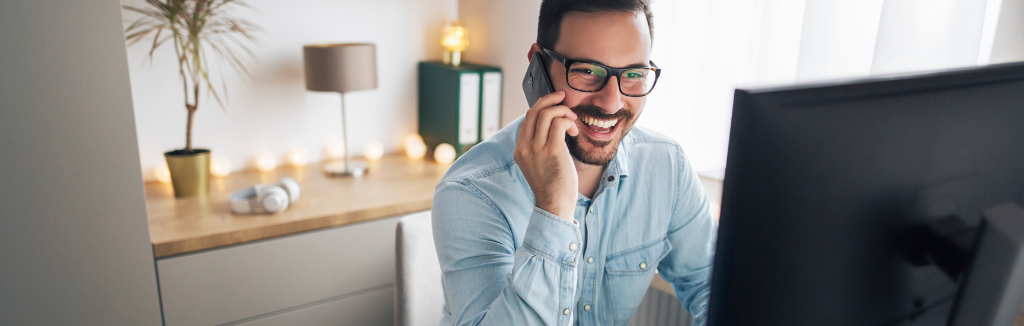 Happy business man working from home, sitting at his desk, in front of his computer monitor, talking on his mobile phone